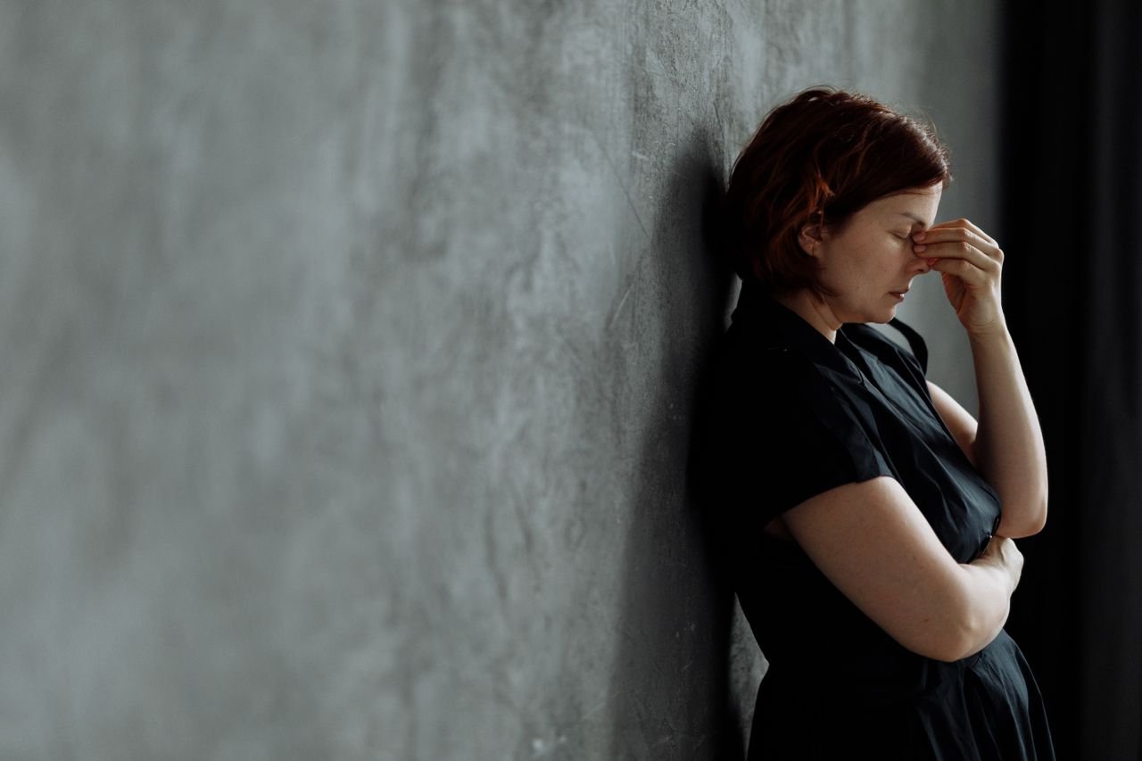 A woman in stress with eyes closed, leaning against a concrete wall indoors.