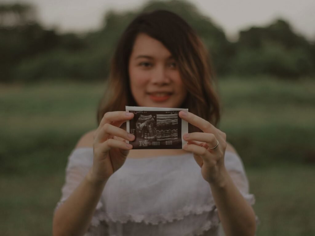 A smiling pregnant woman holds an ultrasound photo outdoors, symbolizing joy and anticipation.