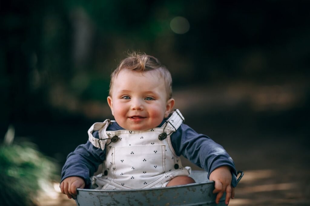 Charming baby in white overalls smiling joyfully while seated outdoors.