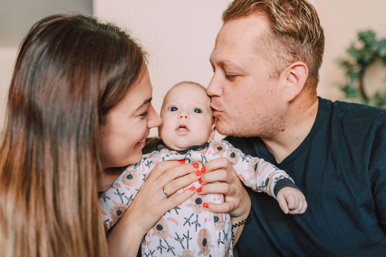 DONATIONS Two parents leaning in to kiss their newborn baby, held between them.