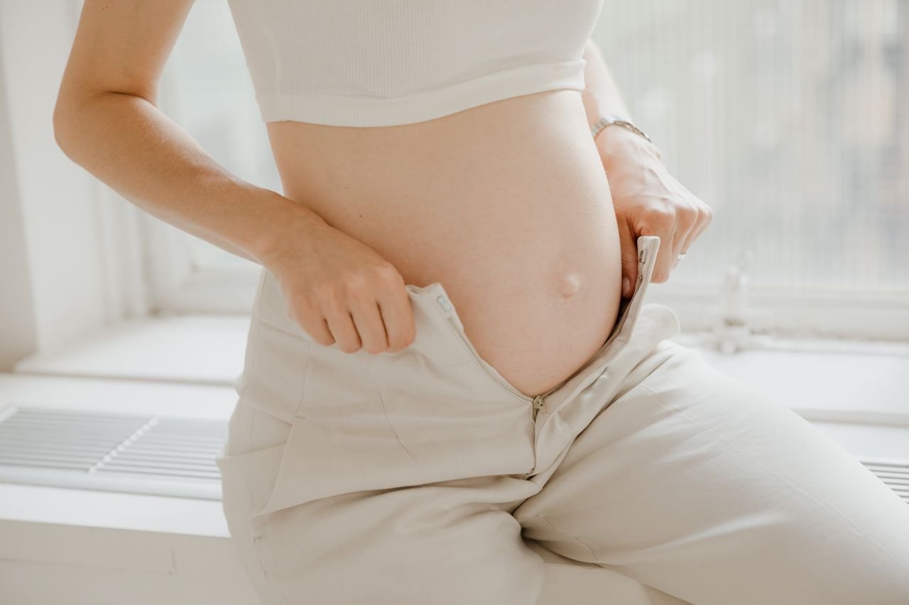 A close-up of a pregnant woman adjusting her pants, highlighting her belly in a sunlit room.